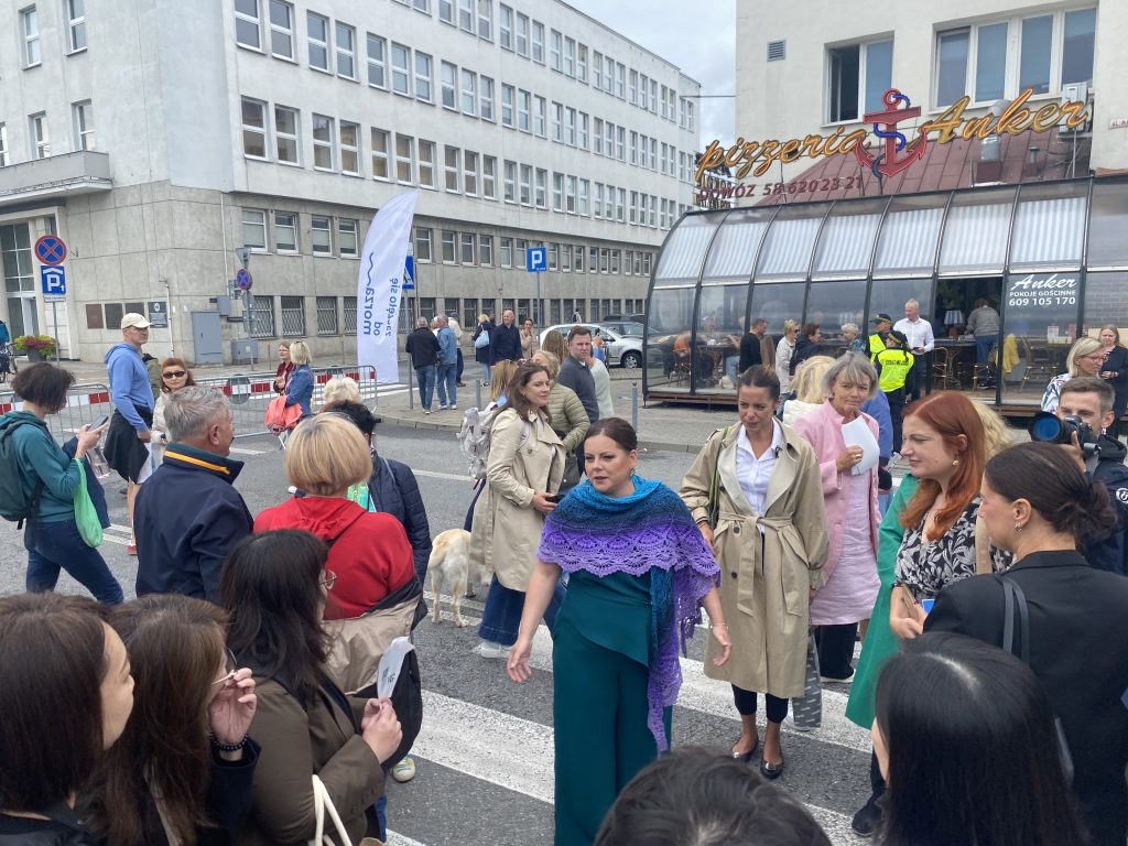 Aleksandra Kosiorek w niebieskiej stylizacji z fioletową chustą rozmawia z ludźmi zgromadzonymi na miejskiej ulicy podczas Gdyńskiego Stołu Urodzinowego