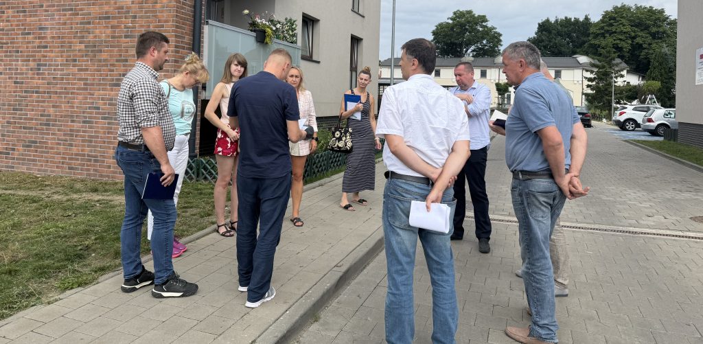 Spotkanie na świeżym powietrzu przed wielorodzinnym budynkiem z ceglaną elewacją. Grupa kilku osób stoi w kręgu na chodniku, rozmawiając i przeglądając trzymane w rękach dokumenty. Spotkanie dotyczyło usterek w nowych blokach komunalnych w Słupsku