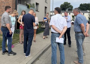 Spotkanie na świeżym powietrzu przed wielorodzinnym budynkiem z ceglaną elewacją. Grupa kilku osób stoi w kręgu na chodniku, rozmawiając i przeglądając trzymane w rękach dokumenty. Spotkanie dotyczyło usterek w nowych blokach komunalnych w Słupsku