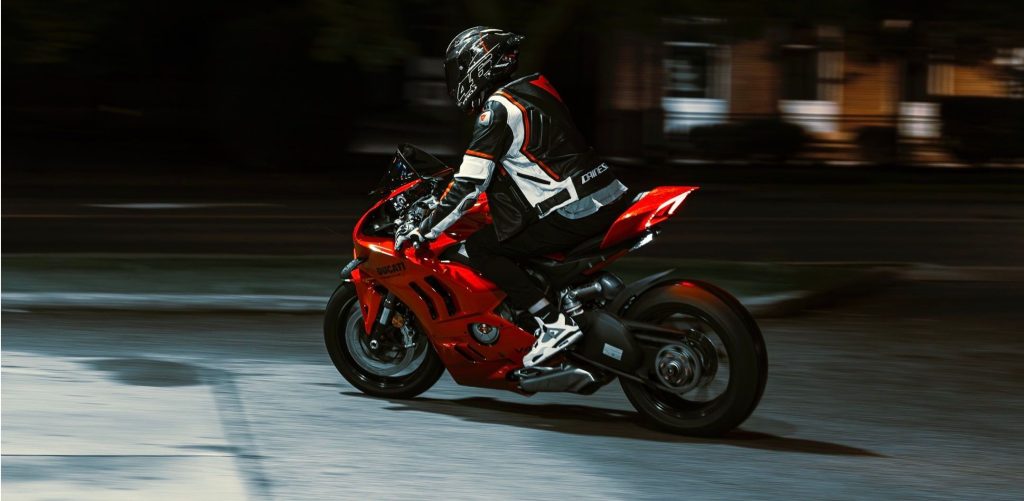 Motocyklista w kasku i skórzanym kombinezonie jedzie nocą na czerwonym motocyklu sportowym po oświetlonej ulicy. Zdjęcie jest ilustracją do artykułu o motocyklach hałasujących nocą w Gdyni