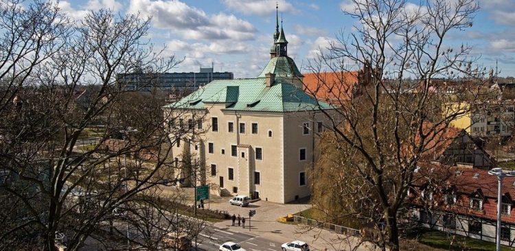 Widok z góry na Zamek Książąt Pomorskich w Słupsku – jasny, kilkupiętrowy budynek z zielonym miedzianym dachem i ozdobną wieżyczką, będący siedzibą Muzeum Pomorza Środkowego, sfotografowany zza koron drzew