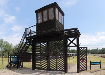 Widok na bramę wejściową i drewnianą wieżę strażniczą na terenie byłego obozu koncentracyjnego Stutthof; wokół widoczne są obozowe baraki i druty kolczaste. Na Cmentarzu Ofiar Marszu Śmierci w Rybnie pochowano szczątki czterech osób - prawdopodobnych ofiar ewakuacji obozu koncentracyjnego KL Stutthof z 1945 roku