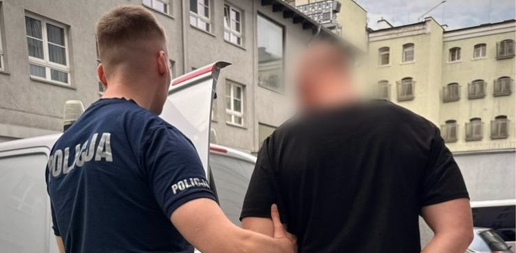 Policjant w granatowej koszulce prowadzi do radiowozu zatrzymanego mężczyznę w czarnej koszulce. Prowadzony jest podejrzany o posiadanie i handel narkotykami. W przyczepie kempingowej na gdańskiej Oruni ukrywał 9 kg narkotyków