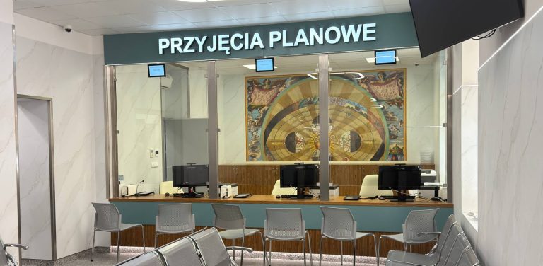 Wnętrze nowo otwartego Punktu Przyjęć Planowych w Szpitalu im. Mikołaja Kopernika w Gdańsku. Nowoczesna lada recepcyjna z przeszklonymi stanowiskami obsługi wyposażonymi w komputery oraz krzesła dla pacjentów na tle ściany z grafiką historyczną