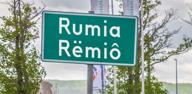 Zielona tablica drogowa z dwujęzyczną nazwą miejscowości: "Rumia" w języku polskim oraz "Rëmiô" w języku kaszubskim, ustawiona na tle drzew i masztów flagowych