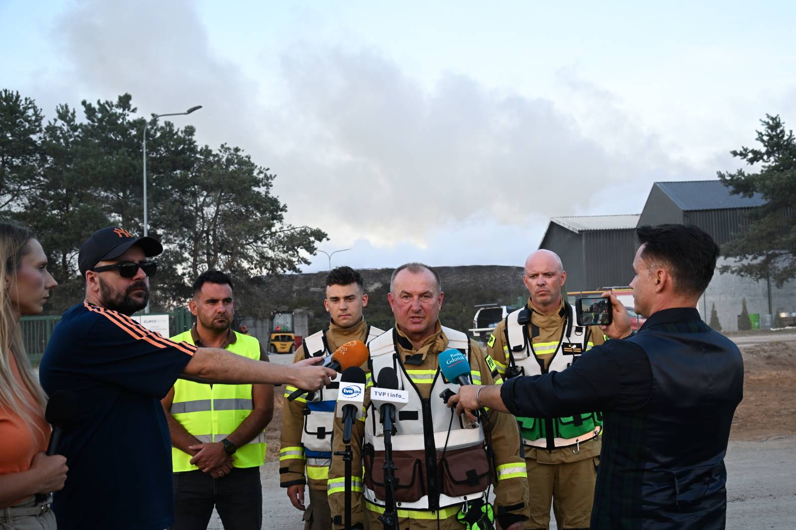 Chlewnica, 15.08.2026. Pomorski Komendant Wojewódzki PSP nadbryg. Jacek Niewęgłowski (C) podczas wypowiedzi dla mediów w miejscu pożaru Zakładu Zagospodarowania Odpadów w Chlewnicy. Ogień objął cztery hale magazynowe o łącznej powierzchni ponad 5 tysięcy metrów kwadratowych oraz hałdę odpadów zajmującą około 2 tysiące metrów kwadratowych