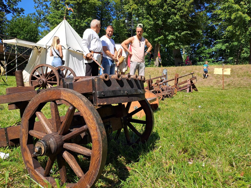 Zamkowy Piknik Historyczny w Człuchowie. Na zielonej trawie stoi drewniane wielolufowe działo (organki) na kołach, a w tle biały namiot obozowy, rekonstruktorzy i turyści w słoneczny dzień