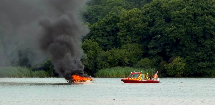 Na jeziorze płonie niewielka łódź motorowa. Z jej pokładu unoszą się wysokie płomienie i gęsty, czarny dym. Obok znajduje się czerwona łódź ratownicza z ratownikami, a w tle widać zalesiony brzeg i trzcinowisko.