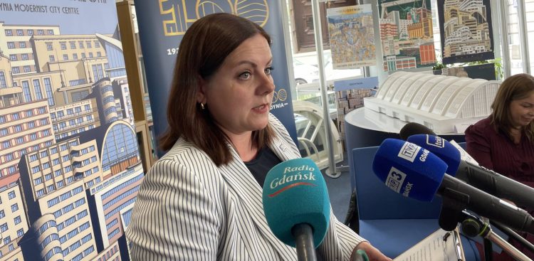 Prezydent Gdyni Aleksandra Kosiorek przemawia do mikrofonów dziennikarzy na konferencji prasowej. W tle znajduje się baner z ilustracjami modernistycznej architektury i napisem "Gdynia Modernist City Centre"oraz makieta budynku