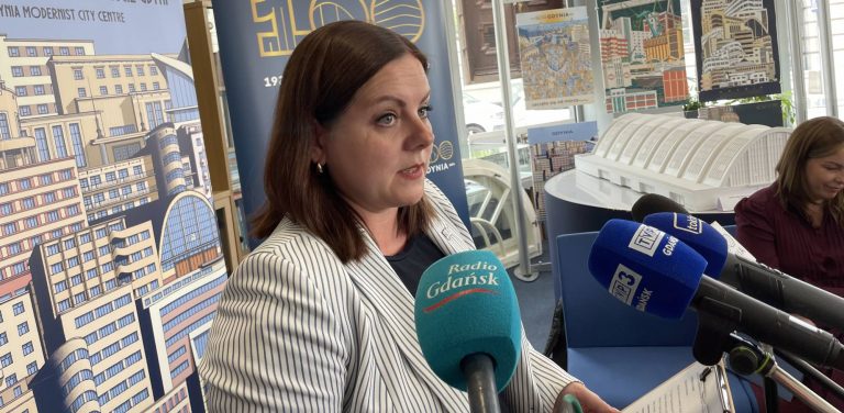 Prezydent Gdyni Aleksandra Kosiorek przemawia do mikrofonów dziennikarzy na konferencji prasowej. W tle znajduje się baner z ilustracjami modernistycznej architektury i napisem "Gdynia Modernist City Centre"oraz makieta budynku