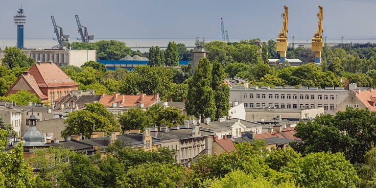 Panorama dzielnicy Nowy Port w Gdańsku z lotu ptaka, ukazująca zwarte zabytkowe kamienice o czerwonych dachach otoczone bujną zielenią drzew. Na dalszym planie widoczne są portowe dźwigi, żurawie, wieża kapitanatu oraz Zatoka Gdańska pod pochmurnym niebem