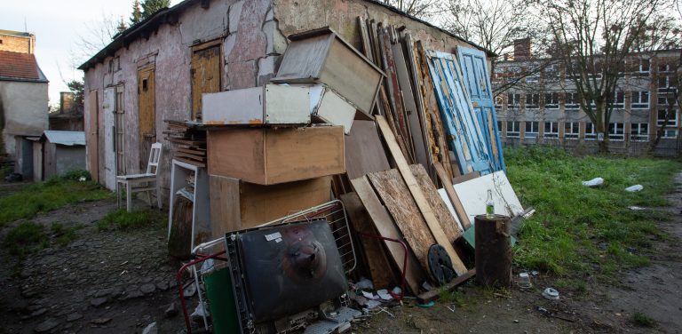Stos starych mebli, płyt drewnianych, starych drzwi i zniszczonego sprzętu AGD porzucony przy ścianie starego budynku