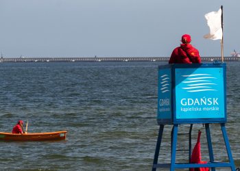 Ratownik w czerwonym ubraniu siedzi na niebieskiej wieży obserwacyjnej z napisem "Gdańsk kąpieliska morskie", nad którą powiewa biała flaga. Obok, na spokojnych falach morza, kołysze się pomarańczowa łódź ratunkowa z drugim ratownikiem. W tle widać falochron. Ratownicy wodni podsumowali sezon 2026