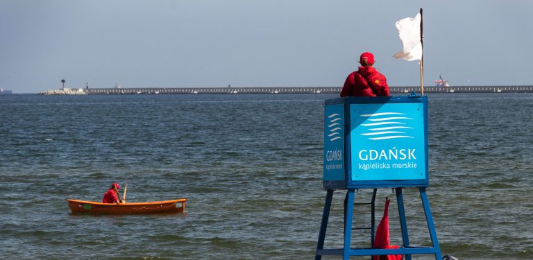 Ratownik w czerwonym ubraniu siedzi na niebieskiej wieży obserwacyjnej z napisem "Gdańsk kąpieliska morskie", nad którą powiewa biała flaga. Obok, na spokojnych falach morza, kołysze się pomarańczowa łódź ratunkowa z drugim ratownikiem. W tle widać falochron. Ratownicy wodni podsumowali sezon 2026