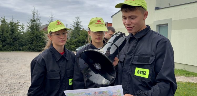 Trójka dzieci z młodzieżowych drużyn pożarniczych. Na pierwszym planie chłopak mówi do megafonu, w ręku trzymając mapę
