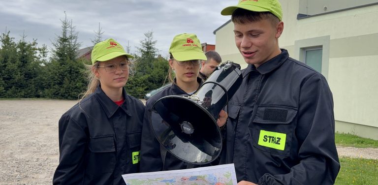 Trójka dzieci z młodzieżowych drużyn pożarniczych. Na pierwszym planie chłopak mówi do megafonu, w ręku trzymając mapę