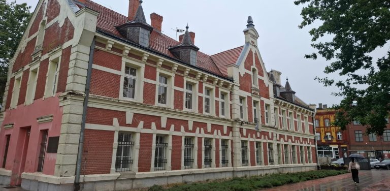 Dwupiętrowy, zabytkowy budynek z czerwonej cegły o neorenesansowej architekturze, z ozdobnymi wieżyczkami i białymi detalami na elewacji. Przed obiektem ciągnie się pas zieleni oraz mokry od deszczu chodnik, po którym idzie osoba z parasolem