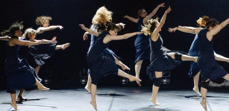 Grupa tancerek w ciemnogranatowych, zwiewnych sukienkach wykonuje dynamiczny układ tańca współczesnego na zaciemnionej scenie teatralnej, zastygając w pełnych ekspresji skokach i wyciągniętych gestach. Trwa nabór do pierwszego Tczewskiego Teatru Tańca
