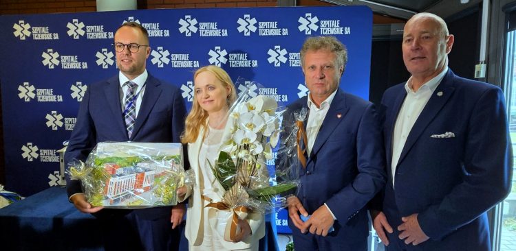 Cztery elegancko ubrane osoby stoją na tle niebieskiej ścianki z logo Szpitali Tczewskich. Mężczyzna po lewej trzyma zapakowaną wizualizację nowego budynku, a kobieta w białym garniturze – bukiet białych storczyków. Placówkę odwiedziła minister zdrowia Jolanta Sobierańska-Grenda (w centrum), która mówiła o rozwoju Szpitali Tczewskich i środkach pozyskanych z Krajowego Planu Odbudowy