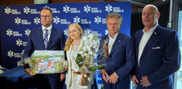 Cztery elegancko ubrane osoby stoją na tle niebieskiej ścianki z logo Szpitali Tczewskich. Mężczyzna po lewej trzyma zapakowaną wizualizację nowego budynku, a kobieta w białym garniturze – bukiet białych storczyków. Placówkę odwiedziła minister zdrowia Jolanta Sobierańska-Grenda (w centrum), która mówiła o rozwoju Szpitali Tczewskich i środkach pozyskanych z Krajowego Planu Odbudowy