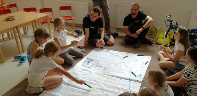 Dzieci i dorośli siedzą na ziemi i rozpisują pomysły na dużym, białym brystolu