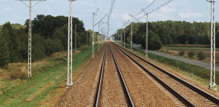 Proste, dwutorowe zelektryfikowane tory kolejowe biegnące przez zielony krajobraz w stronę horyzontu. Po obu stronach torów stoją metalowe słupy trakcyjne.