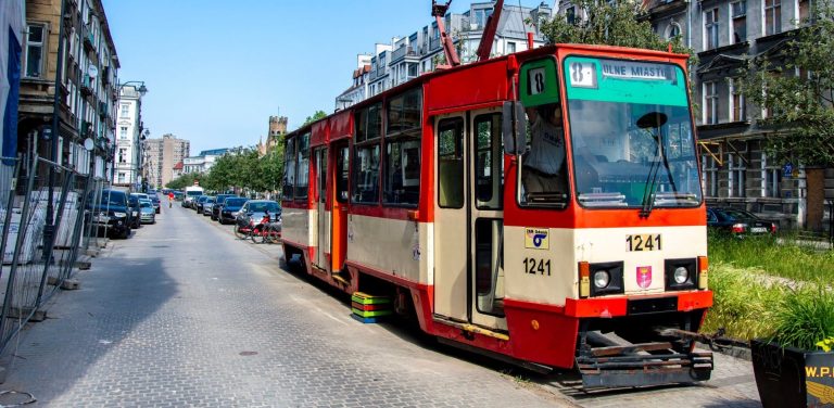 Czerwono-kremowy tramwaj stoi na torach na zabrukowej ulicy w Gdańsku, z tablicą kierunkową „DOLNE MIASTO”, w otoczeniu starych kamienic i zaparkowanych samochodów