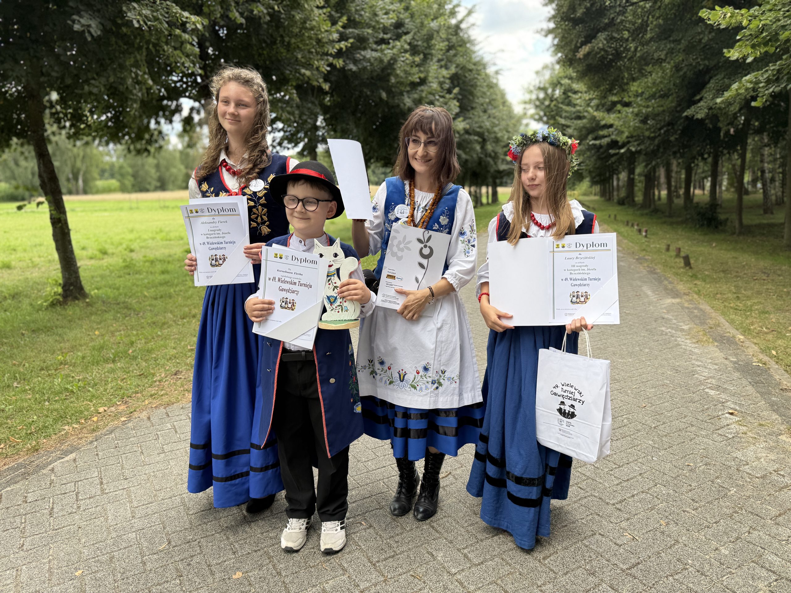 Czworo laureatów i uczestników laureaci 49. Wielewskiego Turnieju Gawędziarzy - trójka dzieci i młodzieży oraz dorosła kobieta - stoi na wybrukowanej alejce w parku. Wszyscy są ubrani w tradycyjne stroje ludowe (niebieskie spódnice, gorsety i kamizelki) i trzymają w rękach dyplomy, statuetkę oraz torbę z upominkami. Dziewczynka po prawej stronie ma na głowie wianek z kolorowych kwiatów