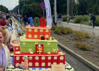 Kobieta stoi obok wielowarstwowej instalacji w kształcie tortu wykonanej na szydełku, ustawionej przy długim stole plenerowym podczas gdyńskiego święta