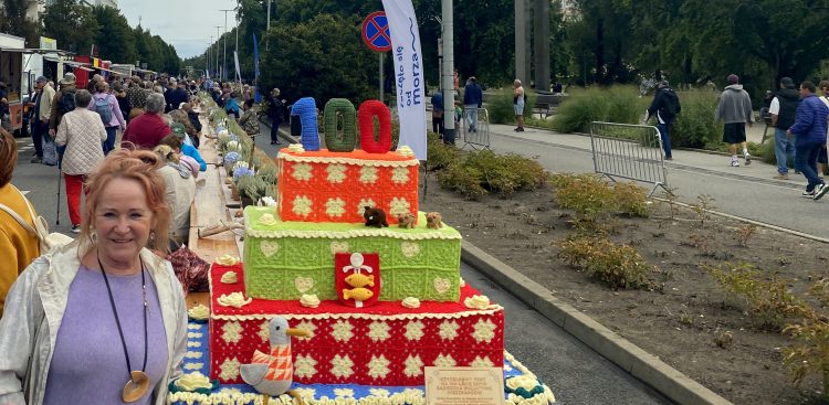 Kobieta stoi obok wielowarstwowej instalacji w kształcie tortu wykonanej na szydełku, ustawionej przy długim stole plenerowym podczas gdyńskiego święta