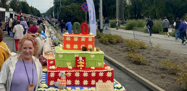 Kobieta stoi obok wielowarstwowej instalacji w kształcie tortu wykonanej na szydełku, ustawionej przy długim stole plenerowym podczas gdyńskiego święta