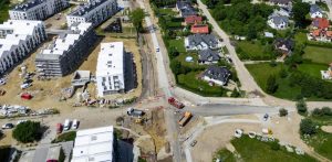 Zdjęcie lotnicze przedstawia trwające prace drogowe i budowlane na skrzyżowaniu ulicy Wiczlińskiej w Gdyni. Widoczny jest głęboki wykop w jezdni, ciężki sprzęt budowlany oraz robotnicy. Po lewej stronie znajduje się duże, nowoczesne osiedle mieszkaniowe w trakcie rozbudowy, a po prawej istniejąca zabudowa jednorodzinna. Wokół wykopu stoją barierki i zaparkowane pojazdy. Całość ilustruje problemy z modernizacją kluczowego odcinka drogi
