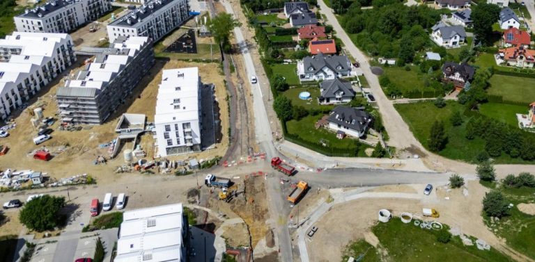 Zdjęcie lotnicze przedstawia trwające prace drogowe i budowlane na skrzyżowaniu ulicy Wiczlińskiej w Gdyni. Widoczny jest głęboki wykop w jezdni, ciężki sprzęt budowlany oraz robotnicy. Po lewej stronie znajduje się duże, nowoczesne osiedle mieszkaniowe w trakcie rozbudowy, a po prawej istniejąca zabudowa jednorodzinna. Wokół wykopu stoją barierki i zaparkowane pojazdy. Całość ilustruje problemy z modernizacją kluczowego odcinka drogi