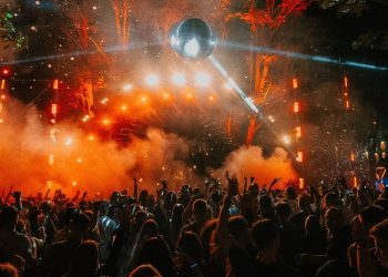 Tłum uczestników bawiących się pod sceną na festiwalu muzycznym w nocy. W tle widoczne są efektowne pomarańczowe światła, dym oraz wielka kula dyskotekowa zawieszona nad publicznością