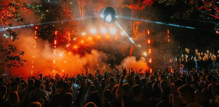 Tłum uczestników bawiących się pod sceną na festiwalu muzycznym w nocy. W tle widoczne są efektowne pomarańczowe światła, dym oraz wielka kula dyskotekowa zawieszona nad publicznością