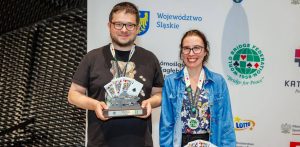 Uśmiechnięte małżeństwo brydżystów, Piotr i Agnieszka Wiankowscy, pozuje na tle ścianki sponsorskiej mistrzostw świata. Mężczyzna trzyma puchar za drugie miejsce z motywem katowickiego Spodka i kart do gry, a kobieta ma na szyi pamiątkowy medal