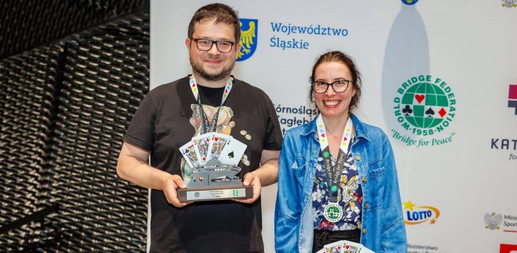 Uśmiechnięte małżeństwo brydżystów, Piotr i Agnieszka Wiankowscy, pozuje na tle ścianki sponsorskiej mistrzostw świata. Mężczyzna trzyma puchar za drugie miejsce z motywem katowickiego Spodka i kart do gry, a kobieta ma na szyi pamiątkowy medal