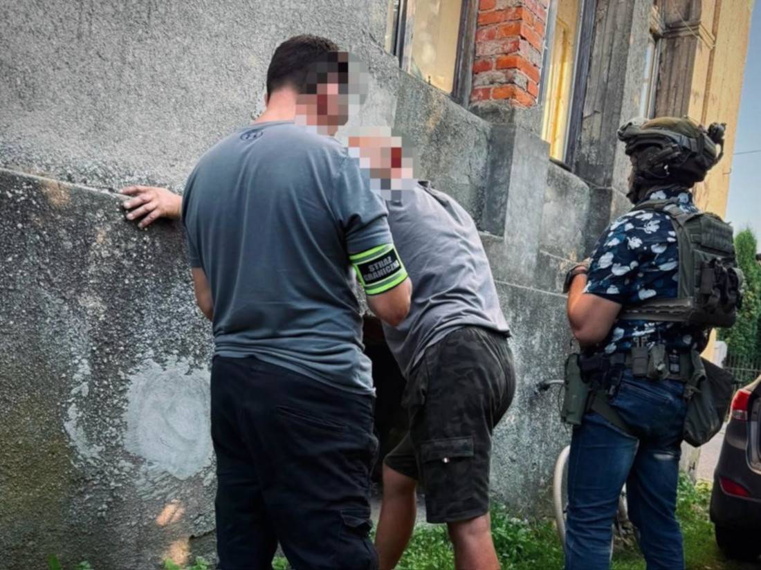 Funkcjonariusz w szarej koszulce z opaską "Straż Graniczna" na ramieniu przyciska do ściany budynku zatrzymanego mężczyznę. Obok nich stoi zamaskowany funkcjonariusz w hełmie i kamizelce taktycznej. Twarze mężczyzn są zamazane
