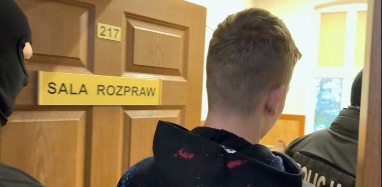 Młody mężczyzna w ciemnej bluzie z kapturem, widziany od tyłu, jest wprowadzany przez zamaskowanych funkcjonariuszy policji do sali rozpraw oznaczonej numerem 217. To podejrzany o brutalne zabójstwo w Słupsku 19-letni Alan G. Zdaniem biegłych w chwili popełnienia zarzucanych mu czynów był w pełni poczytalny