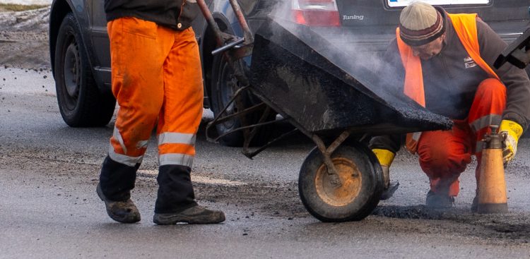 Dwaj drogowcy w pomarańczowych odzieży ostrzegawczej wylewają gorący asfalt z taczki podczas naprawy nawierzchni drogi