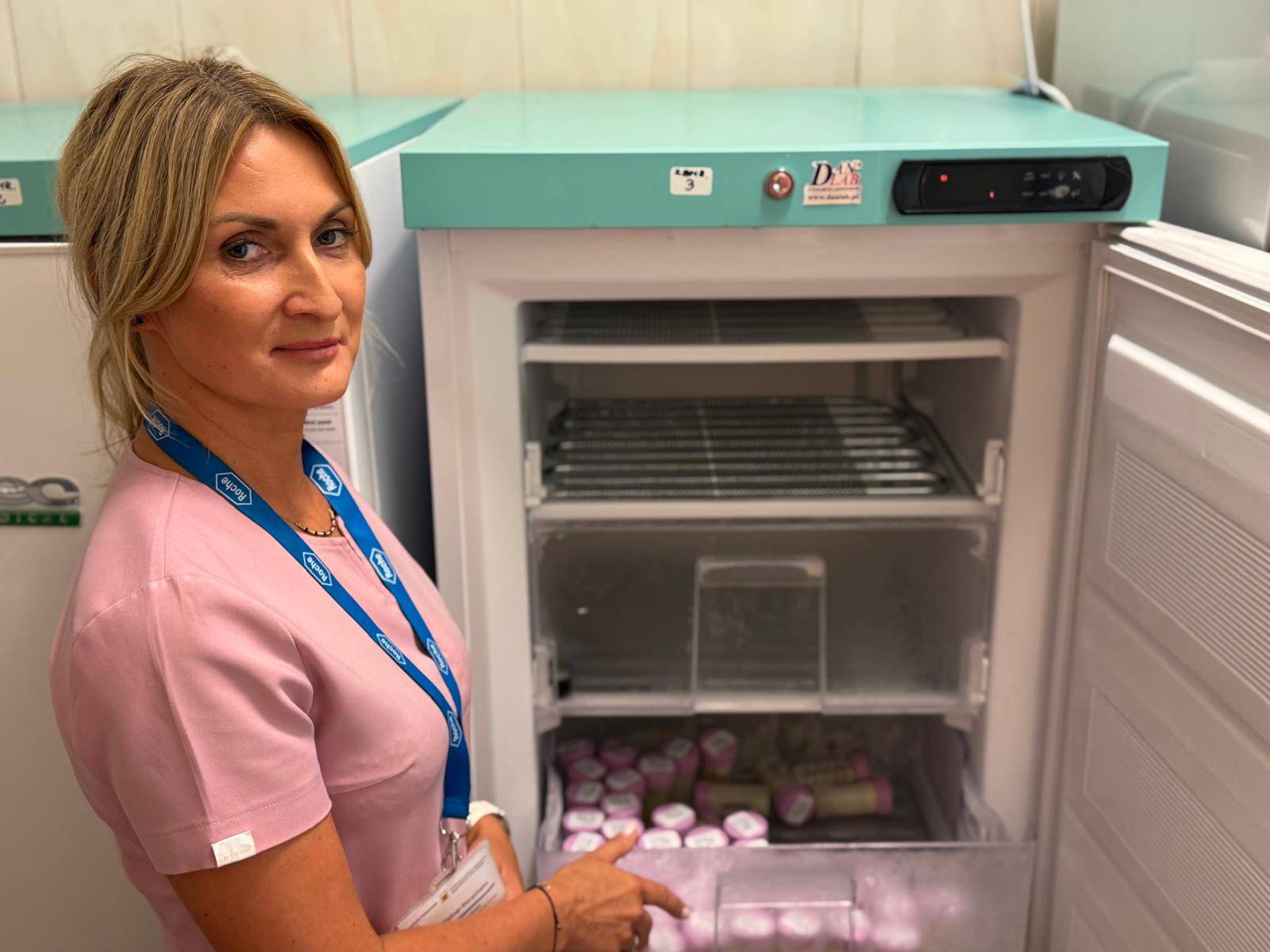 Kobieta w różowym fartuchu medycznym stoi przy otwartej zamrażarce laboratoryjnej i wskazuje dłonią na pojemniki z mlekiem w dolnej szufladzie; górne półki zamrażarki są puste