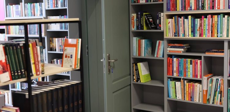 Wnętrze nowoczesnej biblioteki: wysokie, szare regały gęsto zapełnione kolorowymi książkami oraz uchylone drzwi pośrodku