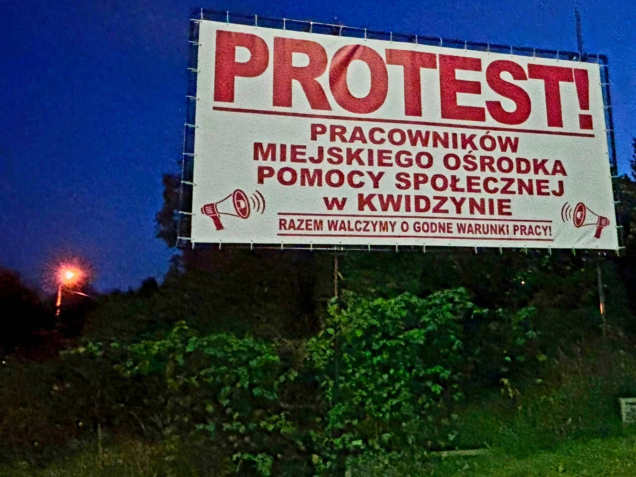 Duży billboard protestacyjny na metalowym stelażu sfotografowany po zmroku na tle ciemnoniebieskiego nieba i zarośli. Na białym tle widnieje wyraźny, czerwony napis: „PROTEST! Pracowników Miejskiego Ośrodka Pomocy Społecznej w Kwidzynie. Razem walczymy o godne warunki pracy!” oraz symbole megafonów. W tle po lewej stronie widać świecącą latarnię uliczną.