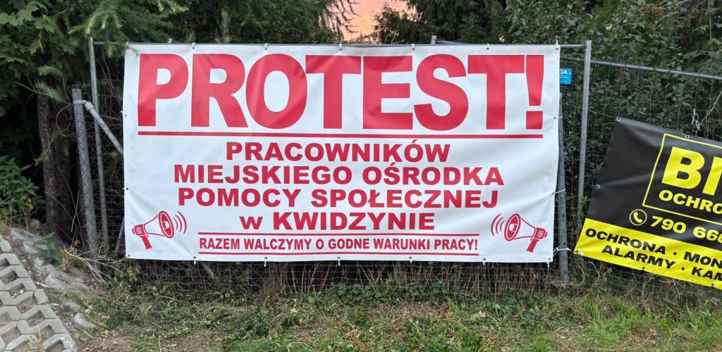 Biały baner protestacyjny zawieszony na ogrodzeniu z czerwonym napisem: „PROTEST! Pracowników Miejskiego Ośrodka Pomocy Społecznej w Kwidzynie. Razem walczymy o godne warunki pracy!” oraz grafikami megafonów po bokach.