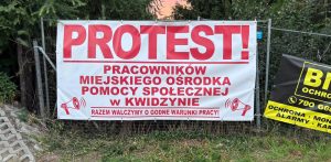Biały baner protestacyjny zawieszony na ogrodzeniu z czerwonym napisem: „PROTEST! Pracowników Miejskiego Ośrodka Pomocy Społecznej w Kwidzynie. Razem walczymy o godne warunki pracy!” oraz grafikami megafonów po bokach.