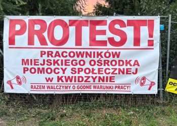 Biały baner protestacyjny zawieszony na ogrodzeniu z czerwonym napisem: „PROTEST! Pracowników Miejskiego Ośrodka Pomocy Społecznej w Kwidzynie. Razem walczymy o godne warunki pracy!” oraz grafikami megafonów po bokach.