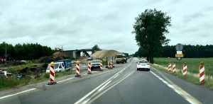 Widok z perspektywy kierowcy na asfaltową drogę ze zwężeniem i biało-czerwonymi słupkami ostrzegawczymi po bokach. Po lewej stronie trwają prace budowlane przy nasypie i wiadukcie, a po prawej widoczny jest żółty znak objazdu oraz zielone pola. Przed jadącym autem porusza się biały samochód osobowy. Przejazd starą drogą krajową nr 6 w powiecie słupskim, w sąsiedztwie budowy nowej trasy ekspresowej. Choć odcinek z tymczasowym oznakowaniem jest w rzeczywistości przejezdny, w systemach nawigacji GPS figuruje jako zamknięty, co powoduje problemy lokalnych przedsiębiorców