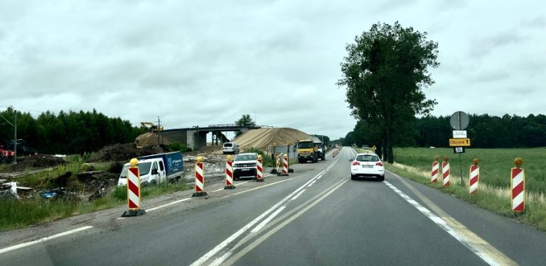 Widok z perspektywy kierowcy na asfaltową drogę ze zwężeniem i biało-czerwonymi słupkami ostrzegawczymi po bokach. Po lewej stronie trwają prace budowlane przy nasypie i wiadukcie, a po prawej widoczny jest żółty znak objazdu oraz zielone pola. Przed jadącym autem porusza się biały samochód osobowy. Przejazd starą drogą krajową nr 6 w powiecie słupskim, w sąsiedztwie budowy nowej trasy ekspresowej. Choć odcinek z tymczasowym oznakowaniem jest w rzeczywistości przejezdny, w systemach nawigacji GPS figuruje jako zamknięty, co powoduje problemy lokalnych przedsiębiorców