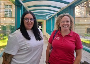 Dwie uśmiechnięte żeglarki, Emilia Witkowska w białej koszulce i okularach oraz Anna Majewska w różowej koszulce polo klubu Sailing Factory, stojące w przeszklonym łączniku budynku Radia Gdańsk
