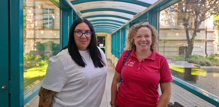 Dwie uśmiechnięte żeglarki, Emilia Witkowska w białej koszulce i okularach oraz Anna Majewska w różowej koszulce polo klubu Sailing Factory, stojące w przeszklonym łączniku budynku Radia Gdańsk
